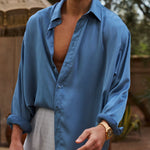Man wearing a blue shirt and sunglasses outdoors with trees and an umbrella in the background