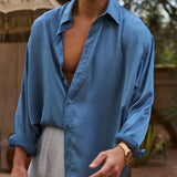 Man wearing a blue shirt and sunglasses outdoors with trees and an umbrella in the background