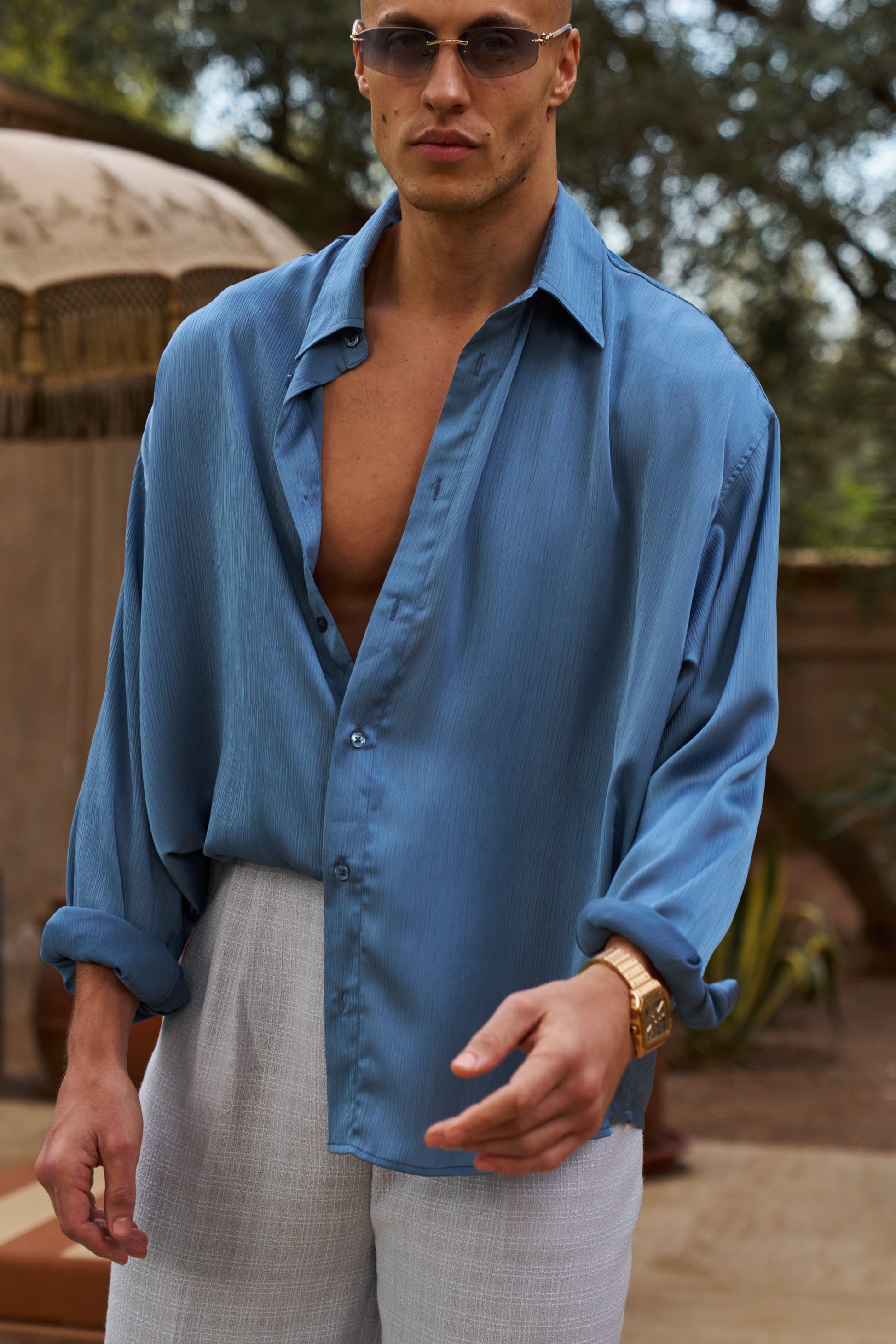 Man wearing a blue shirt and sunglasses outdoors with trees and an umbrella in the background